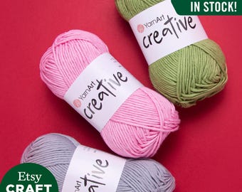 May include: Three balls of yarn in pink, green, and gray. Each ball has a white label with the text "YarnArt creative" on it. The background is red. The text "ALL COLORS IN STOCK!" is in a white box with a black border. The text "Etsy CRAFT MAXI SHOP" is in a green circle.