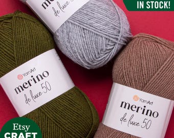 Pode incluir: Três novelos de fio YarnArt merino de luxe 50 em cinza, verde e marrom. Os novelos estão dispostos sobre um fundo vermelho. O texto "TODAS AS CORES EM ESTOQUE!" é visível no canto superior direito da imagem.