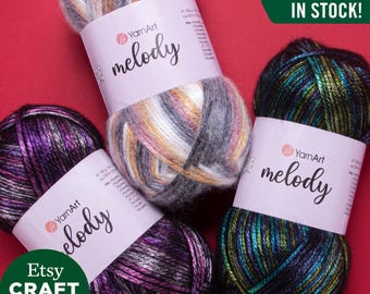 Op de afbeelding: Drie bollen YarnArt Melody garen in verschillende kleurcombinaties. Het garen is een zachte, pluche mix van vezels met een subtiele glans. De kleuren zijn een mix van zwart, wit, goud, roze, groen en blauw. De bollen zijn gelabeld met "YarnArt Melody" en "Alle kleuren op voorraad!"