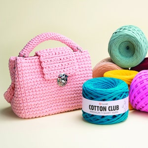 May include: A pink crocheted handbag with a silver clasp and a handle. The bag is sitting on a light yellow surface next to several balls of yarn in different colors, including teal, yellow, pink, and green. The yarn is labeled "Cotton Club Premium T-Shirt Yarn".