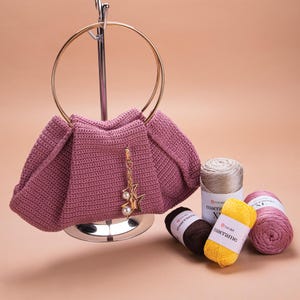 May include: A pink crocheted handbag with a gold ring handle and a star-shaped charm. The bag is displayed on a metal stand with three spools of yarn in beige, yellow, and pink.