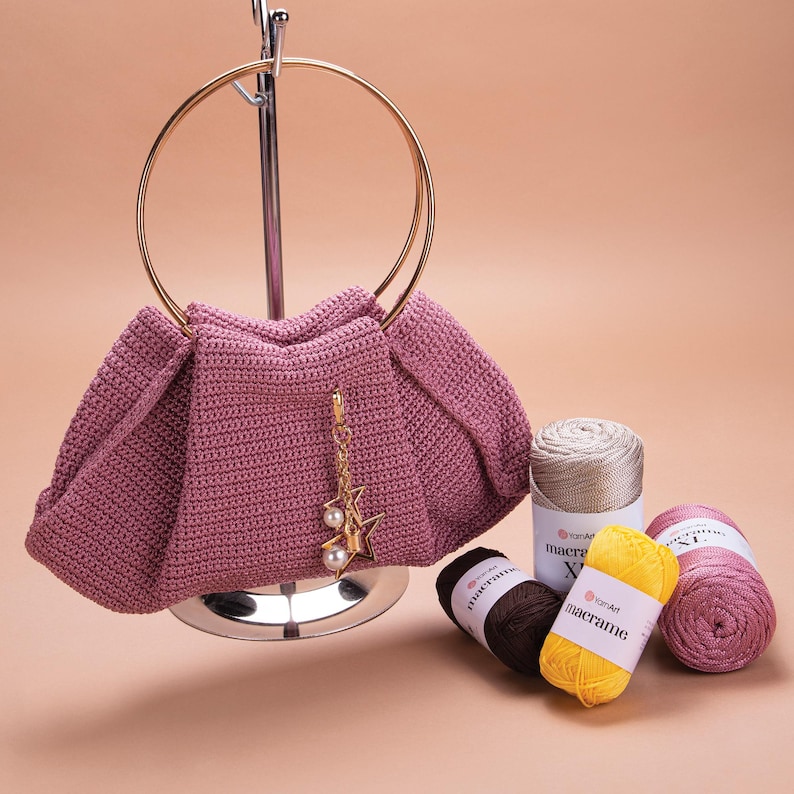 May include: A pink crocheted handbag with a gold ring handle and a star-shaped charm. The bag is displayed on a metal stand with three spools of yarn in beige, yellow, and pink.