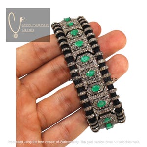 Può includere: Un bracciale nero e argento con pietre preziose color smeraldo. Il bracciale presenta una fascia nera intrecciata con maglie argentate di forma geometrica. Le maglie sono adornate da piccole pietre trasparenti e gemme verdi ovali.