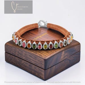 May include: A bracelet featuring a brown braided cord band with a silver-toned clasp. The bracelet is adorned with a series of oval-shaped multi-colored gemstones, each surrounded by a halo of small clear stones, arranged along the cord.