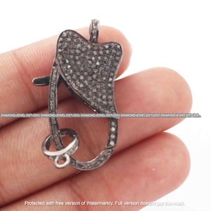 Op de afbeelding: Een donkergrijze, met diamanten bezette sieradensluiting. De sluiting heeft een unieke vorm met een hartvormige bovenkant en een scharniersluiting. De sluiting wordt in een hand gehouden, wat het ingewikkelde ontwerp en de sprankelende details laat zien.