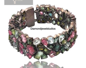 May include: A silver-toned bracelet with a variety of colorful gemstones. The bracelet is encrusted with a mix of pink, green, blue, and clear stones. The clasp is visible, and the text "Diamondjewelstudios" is present.