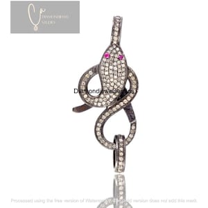 May include: A snake-shaped pendant with a clasp, crafted from dark metal and encrusted with small, sparkling stones. The snake's head features two small, red gemstones for eyes. The pendant is designed to be a jewelry clasp.