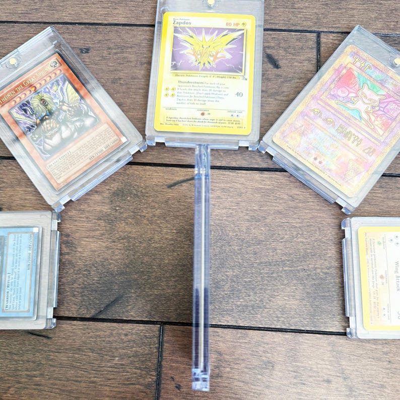 May include: A collection of trading cards, including a Zapdos Pok&eacute;mon card, displayed in clear protective cases. The cards are arranged on a wooden surface, showcasing various designs and colors.
