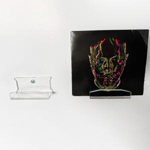 May include: A black album cover with a colorful, abstract face design is displayed on a clear acrylic stand. A second clear acrylic shelf is to the left. The album art features shades of green, pink, and yellow.