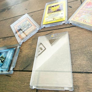 May include: Several clear plastic card holders with various trading cards inside. The cards feature colorful illustrations and text. The holders are arranged on a wooden surface, showcasing the cards' designs and protective cases.
