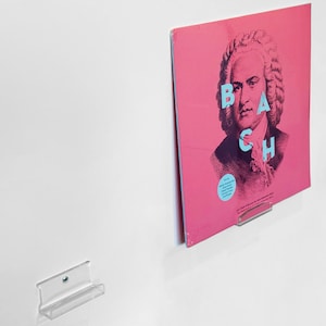 May include: A pink vinyl record album cover featuring a portrait of Johann Sebastian Bach with the name "BACH" in turquoise letters. The album is displayed on a clear, wall-mounted record holder.