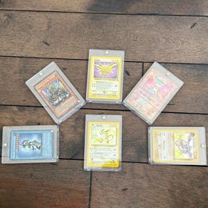 May include: Six collectible trading cards in protective sleeves. The cards feature various characters and designs, including a yellow Raichu, a purple Zapdos, and a blue monster. The cards are arranged on a wooden surface.