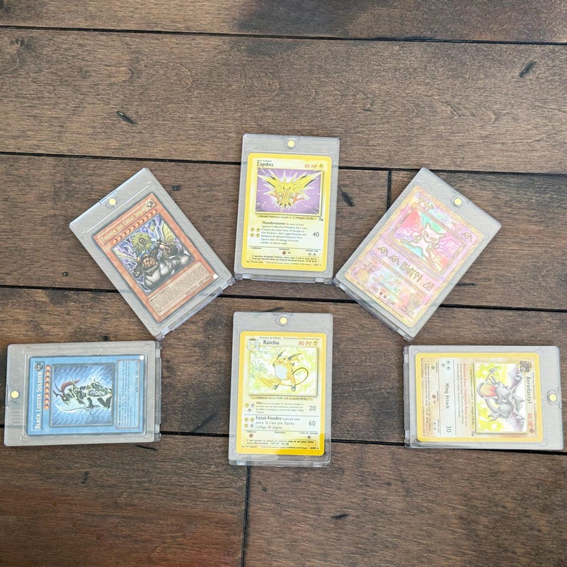 May include: Six collectible trading cards in protective sleeves. The cards feature various characters and designs, including a yellow Raichu, a purple Zapdos, and a blue monster. The cards are arranged on a wooden surface.