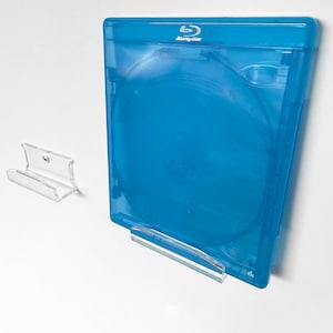 Puede incluir: Un soporte de pared transparente de acrílico para una caja de disco Blu-ray. La caja es de color azul vibrante y presenta el logotipo de Blu-ray Disc. El soporte está diseñado para sujetar la caja de forma segura, mostrando el disco.