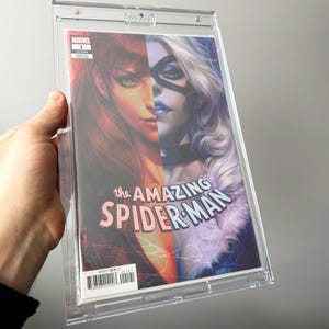 Puede incluir: Un cómic titulado "The Amazing Spider-Man" en una funda protectora transparente. La portada presenta dos rostros femeninos, uno con cabello rojo y otro con cabello blanco y una máscara negra. El logotipo de Marvel es visible.