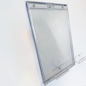 Puede incluir: Un portacarteles de acrílico transparente de forma rectangular. Montado en una pared blanca, presenta un aspecto claro y transparente. Cuenta con soportes superior e inferior para sujetar un letrero o documento. Diseñado para la exhibición de información.