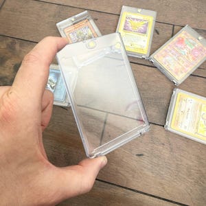 May include: Clear plastic card holders with various trading cards inside. The holders are rectangular and have a snap closure. Several cards are visible, featuring colorful illustrations and text. The cards appear to be collectible items.
