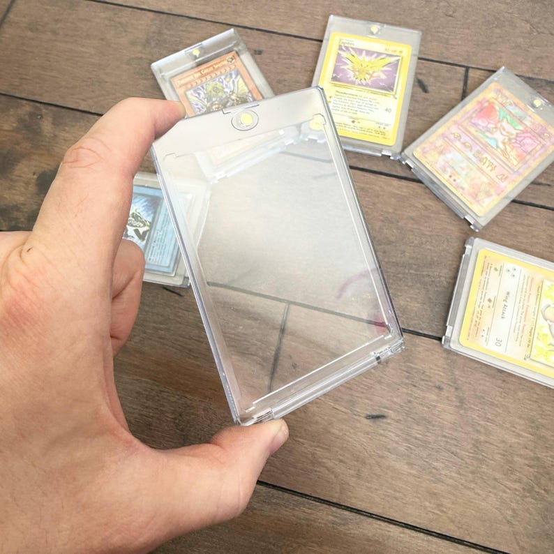May include: Clear plastic card holders with various trading cards inside. The holders are rectangular and have a snap closure. Several cards are visible, featuring colorful illustrations and text. The cards appear to be collectible items.