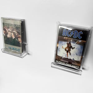May include: Two cassette tapes in clear plastic cases, displayed on clear stands. One features "The Beatles" album art, the other "AC/DC" with the text "BLOW UP YOUR VIDEO". The tapes are set against a white background.