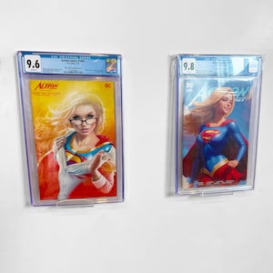 Puede incluir: Dos cómics de Supergirl, calificados y enmarcados. El cómic de la izquierda, Action Comics #1066, está calificado con 9.6, con Supergirl con gafas. El cómic de la derecha está calificado con 9.8, con Supergirl con su traje clásico. Fondo blanco.