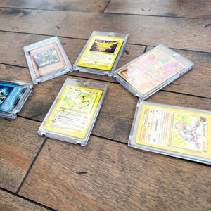 May include: Several trading cards in clear plastic cases are displayed on a wooden surface. The cards feature various colorful illustrations and text, showcasing different characters and designs. The cards are in protective sleeves.