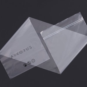 May include: Two transparent plastic bags, one overlapping the other, against a dark background. The bag on the left has the word "ARKORUS" printed in black, along with three small circular symbols. The other bag has a white zipper closure.