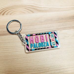 May include: A clear acrylic keychain with a pink, blue and black striped design. The keychain features the text "HOBI PALOOZA 07.31.22".