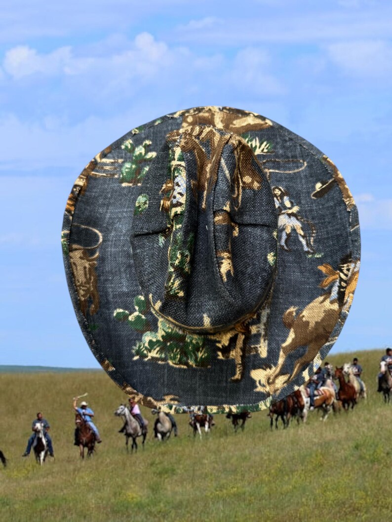 Puede incluir: Un sombrero de vaquero con una base de denim negro y un estampado colorido de vaqueros montando a caballo. El estampado presenta una gama de colores verde, marr&oacute;n y amarillo.