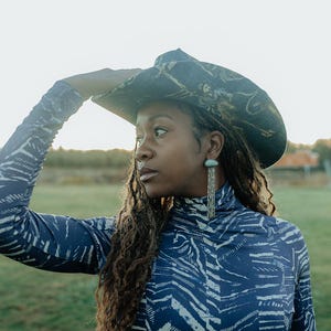 Puede incluir: Una persona lleva una camisa de cuello alto con estampado azul y blanco con un sombrero de vaquero con estampado negro y dorado. Lleva pendientes largos de plata.