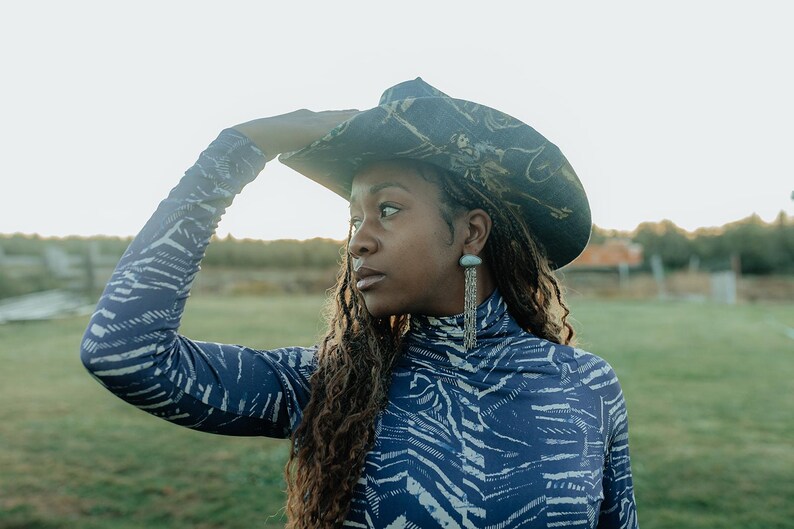 Puede incluir: Una persona lleva una camisa de cuello alto con estampado azul y blanco con un sombrero de vaquero con estampado negro y dorado. Lleva pendientes largos de plata.