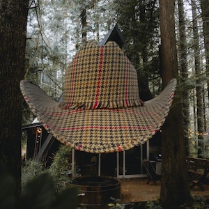 Puede incluir: Un sombrero vaquero de cuadros marrones y beige con ribete rojo y blanco. El sombrero flota frente a una cabaña de madera en un bosque.