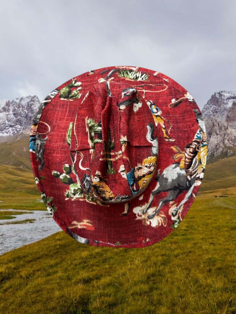 Puede incluir: Un sombrero de vaquero rojo con un estampado de tem&aacute;tica western que presenta vaqueros, caballos y cactus. El sombrero est&aacute; hecho de una tela tejida y tiene un ala ancha.