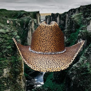 Puede incluir: Un sombrero vaquero de estampado de leopardo marrón y negro con ribete de flecos. El sombrero flota en el aire entre dos acantilados rocosos con un arroyo que corre entre ellos.