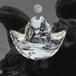 Puede incluir: Un sombrero vaquero blanco con un estampado en blanco y negro de una escena con tres personas. El sombrero tiene un ala ancha y una corona puntiaguda.