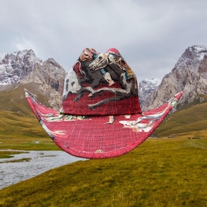Puede incluir: Un sombrero vaquero rojo con un estampado colorido de una escena con caballos y jinetes. El sombrero flota frente a una cordillera con picos nevados.