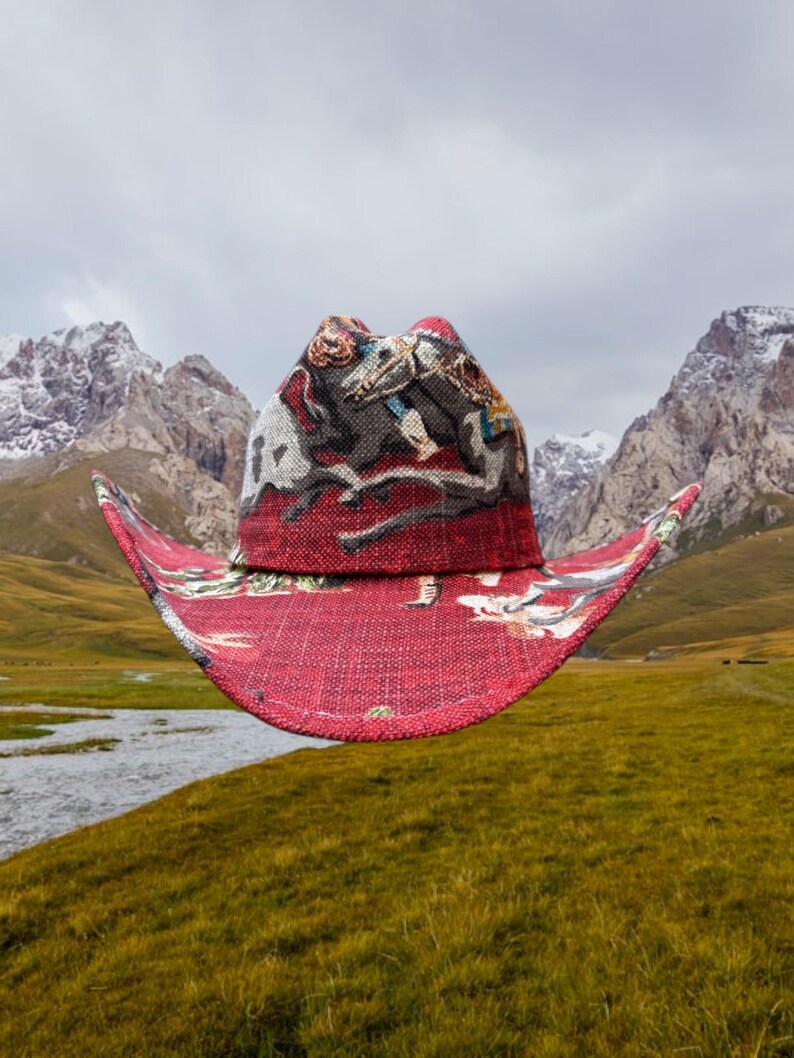 Puede incluir: Un sombrero vaquero rojo con un estampado colorido de una escena con caballos y jinetes. El sombrero flota frente a una cordillera con picos nevados.