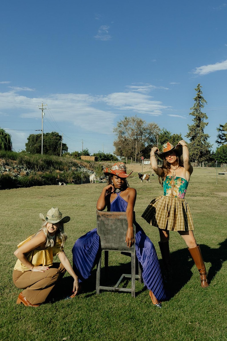 Puede incluir: Tres mujeres con sombreros de vaquero posan al aire libre. Una mujer est&aacute; sentada en una silla de madera con un vestido morado y un sombrero estampado en rojo, blanco y azul. Otra mujer est&aacute; de pie con una blusa estampada en verde y dorado, una falda de cuadros marrones y botas marrones. La tercera mujer lleva una blusa amarilla, pantalones marrones y un sombrero de vaquero marr&oacute;n.