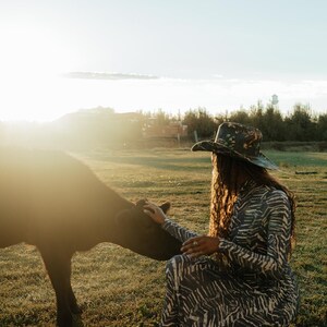 Puede incluir: Una mujer con un sombrero de vaquero con estampado floral y un vestido de estampado de cebra est&aacute; acariciando un ternero negro en un campo. El sol brilla intensamente en el fondo.
