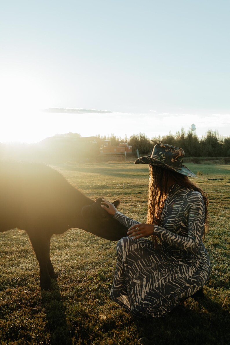 Puede incluir: Una mujer con un sombrero de vaquero con estampado floral y un vestido de estampado de cebra est&aacute; acariciando un ternero negro en un campo. El sol brilla intensamente en el fondo.