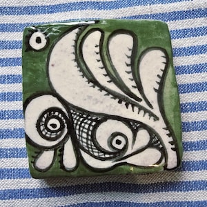 Könnte beinhalten: Quadratische Keramikfliese mit grünem Hintergrund und einem weißen und schwarzen abstrakten Design. Das Design zeigt wirbelnde Formen und ein fischähnliches Motiv. Die Fliese liegt auf einem blau-weiß gestreiften Stoff.