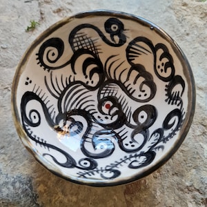 Könnte beinhalten: Eine weiße Keramikschüssel mit einem abstrakten Schwarz-Weiß-Design. Das Design zeigt wirbelnde Linien und Formen, die ein Gefühl von Bewegung und Energie erzeugen. Die Schüssel hat eine glänzende Oberfläche und einen dunkelbraunen Rand.