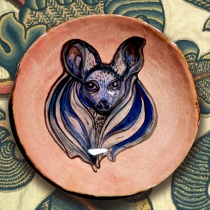 Peut inclure: Assiette ronde en céramique peinte à la main, ornée d'une illustration d'un renard à oreilles de chauve-souris. Le fond est rose et le renard est représenté en bleu, gris et blanc. La glaçure de l'assiette présente un effet craquelé.