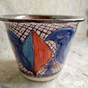May include: A ceramic planter with a white background and a blue, red, and teal floral design. The planter has a black and white grid pattern around the rim.