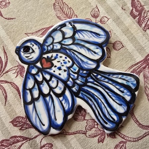 Könnte beinhalten: Keramik-Vogelfigur mit blau-weißem Design. Der Vogel hat ein Herz auf der Brust und schwarze Umrisse. Die Flügel sind ausgebreitet, auf einem gemusterten Stoff.