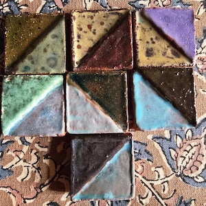 Könnte beinhalten: Eine Sammlung handgefertigter Keramikfliesen, jede quadratisch mit einer diagonalen Farbteilung. Die Fliesen weisen eine Vielzahl von Farben auf, darunter Grün, Braun, Blau und Lila. Die Fliesen sind auf einem gemusterten Stoff angeordnet.
