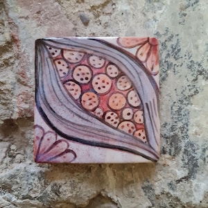 Könnte beinhalten: Quadratische Keramikfliese mit handgemaltem Design. Die Fliese zeigt eine zentrale ovale Form, die mit kleinen, gepunkteten Kreisen in Orange- und Rosatönen gefüllt ist. Das Oval wird von einem grauen und violetten Rand umrahmt, mit floralen Akzenten in den Ecken.