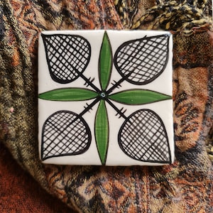 Könnte beinhalten: Eine weiße Keramikfliese mit einem geometrischen Muster in Schwarz und Grün. Das Muster zeigt vier stilisierte Blätter mit einem schwarzen Gittermuster und grünen Stielen.