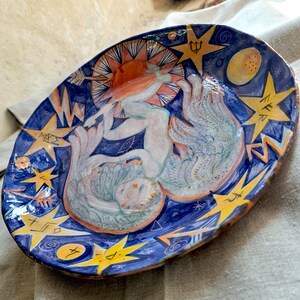 Könnte beinhalten: Ein Keramikteller mit blauem Hintergrund und gelben Sternen. Auf dem Teller ist eine weiße Figur mit Flügeln gemalt, umgeben von Sternen und anderen Symbolen. Der Teller ist mit einer Sonne, einem Mond und einem Dreizack verziert.