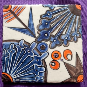 Könnte beinhalten: Eine weiße Keramikfliese mit einem blauen und braunen Blumendesign. Die Fliese hat einen weißen Hintergrund mit blauen und braunen Blumen und Blättern. Die Blumen haben weiße Zentren und sind von blauen Blütenblättern umgeben. Die Blätter sind braun und haben einen weißen Umriss.