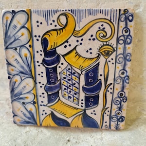 Könnte beinhalten: Eine quadratische Keramikfliese mit einem blauen und gelben Design. Die Fliese zeigt einen stilisierten Buchstaben "D" mit einer dekorativen Verzierung und einem Auge.